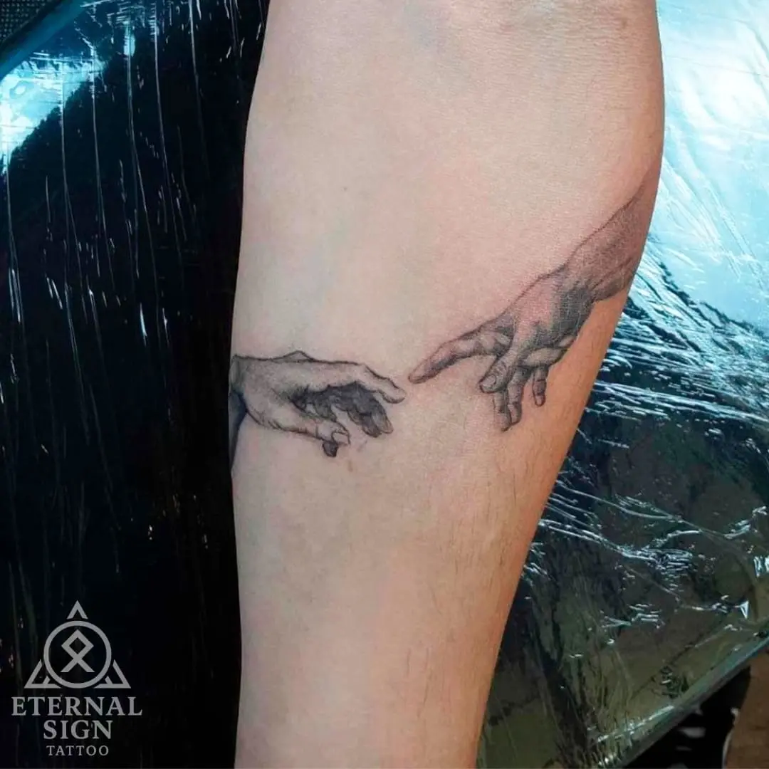 Tatovering av to hender som strekker seg mot hverandre, inspirert av Michelangelos 'Adams skapelse', utført i black & grey stil på underarmen.